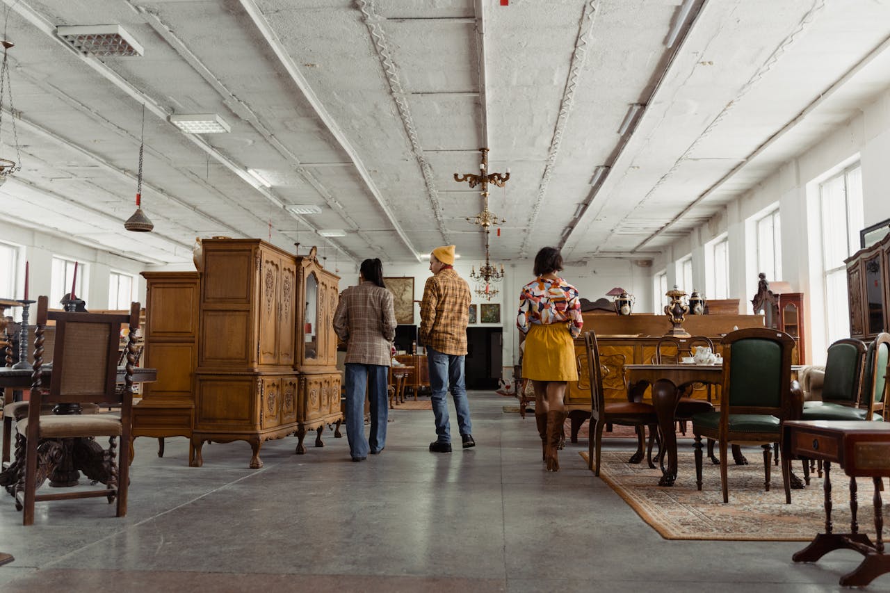 Home Three people browsing vintage wooden furniture in a spacious store interior with high ceilings.