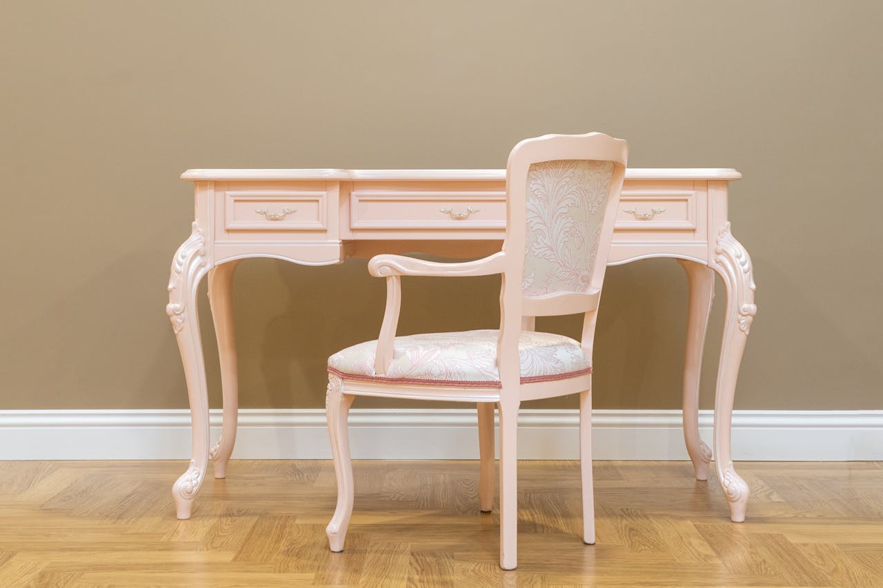 A classic pink vintage desk and matching chair set on a wooden floor with beige wall, perfect for elegant interiors.